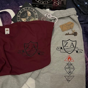 May include: A maroon t-shirt and grey sweatpants with matching black graphic designs. The t-shirt features a crest with "ZA" and "AQUA TERRA IGNIS AER". The sweatpants have the same crest and elemental symbols.