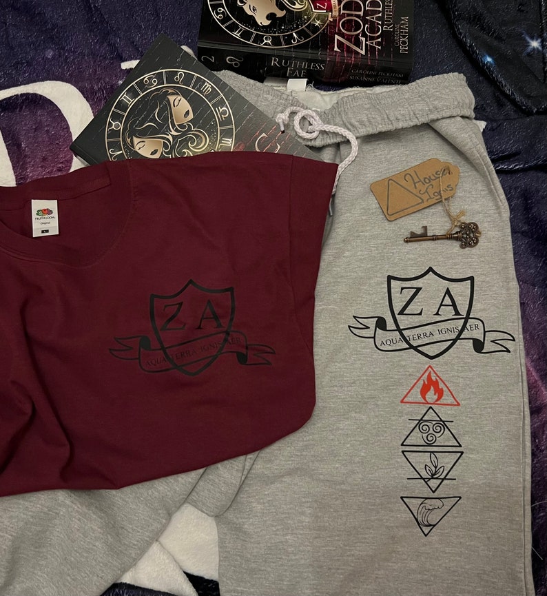 May include: A maroon t-shirt and grey sweatpants with matching black graphic designs. The t-shirt features a crest with "ZA" and "AQUA TERRA IGNIS AER". The sweatpants have the same crest and elemental symbols.