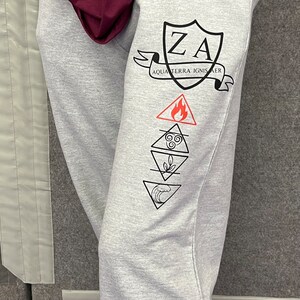 May include: Pale grey sweatpants featuring black graphics. The design includes a shield with "ZA" and the words "AQUA TERRA IGNIS AER" on a banner. Below are four elemental symbols: fire, air, earth, and water. A maroon hoodie is visible at the top.