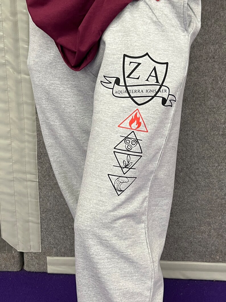 May include: Pale grey sweatpants featuring black graphics. The design includes a shield with "ZA" and the words "AQUA TERRA IGNIS AER" on a banner. Below are four elemental symbols: fire, air, earth, and water. A maroon hoodie is visible at the top.