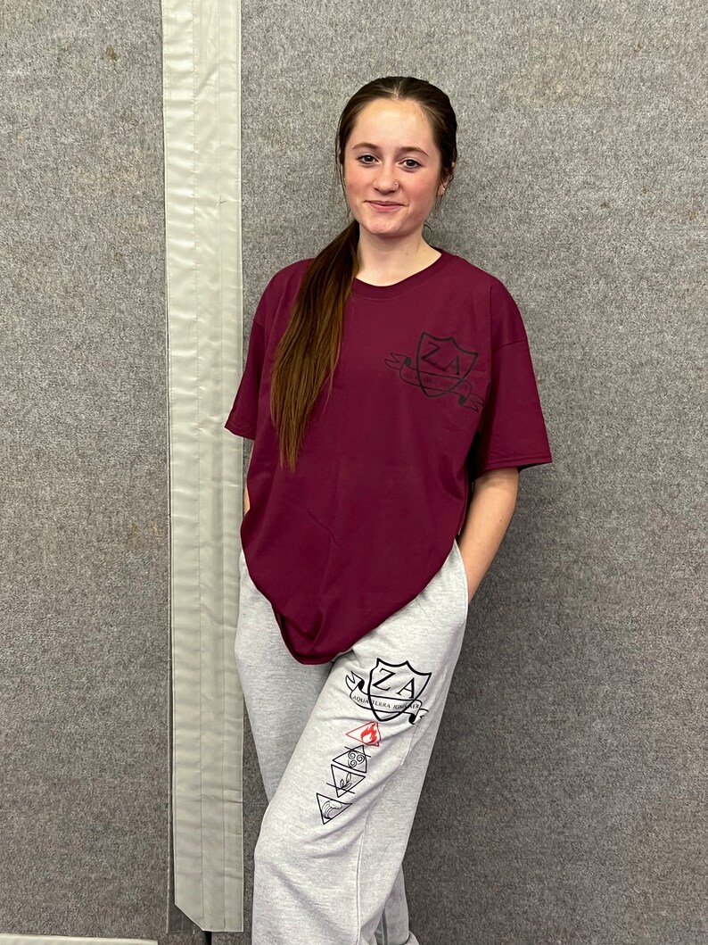 May include: A maroon t-shirt and grey sweatpants. The t-shirt features a shield design with the letters "ZA". The sweatpants have a matching shield design and elemental symbols. The outfit is casual and comfortable, suitable for everyday wear.