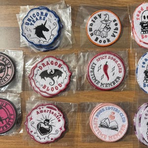 May include: A collection of circular and oval embroidered patches in individual plastic sleeves. Designs include a unicorn, ghosts reading books, a dragon, a chilli pepper, a potion bottle, an open book, and silhouettes with text.
