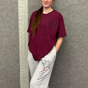 May include: A maroon t-shirt and grey sweatpants. The t-shirt features a black crest design with the letters "ZA". The sweatpants have a matching design on the leg. The outfit is casual and comfortable.