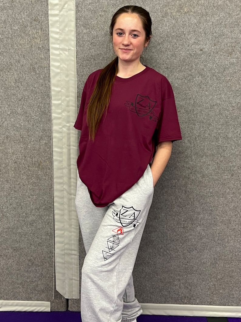 May include: A maroon t-shirt and grey sweatpants. The t-shirt features a black crest design with the letters "ZA". The sweatpants have a matching design on the leg. The outfit is casual and comfortable.