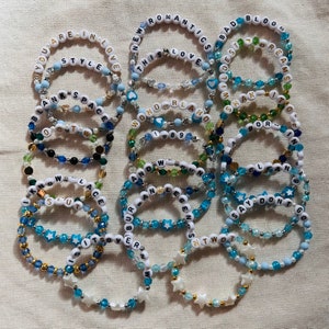 May include: A collection of 15 beaded bracelets with various words and phrases, including "You Are In Love", "New Romantics", "This Love", "Bad Blood", "Blank Space", "Wonderland", "I Know", "Lace", "Burban Legends", "Say Don't Go", and "NTWDT". The bracelets are made with blue, white, and gold beads.