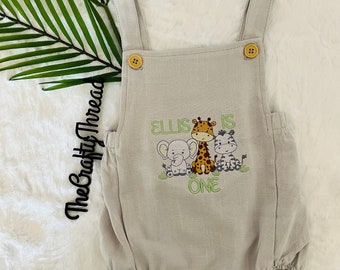 May include: A beige baby onesie with a green embroidered design featuring a giraffe, zebra, and elephant. The text "Ellis is one" is embroidered on the onesie.