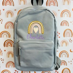May include: Gray backpack with a rainbow and heart embroidery. The backpack has a zippered pocket and adjustable straps. The name "Charlotte" is embroidered below the rainbow.