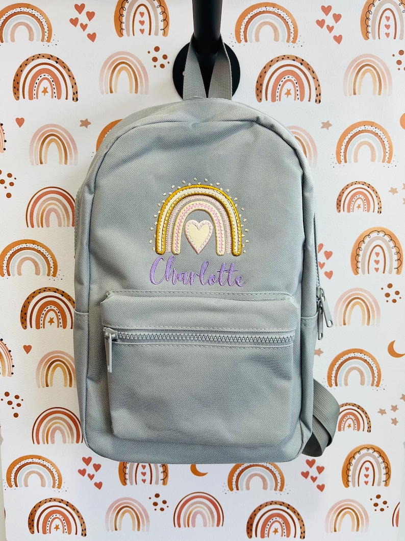 May include: Gray backpack with a rainbow and heart embroidery. The backpack has a zippered pocket and adjustable straps. The name "Charlotte" is embroidered below the rainbow.