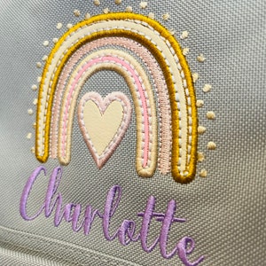 May include: A gray canvas backpack with a rainbow and heart embroidered design. The rainbow is in shades of yellow, pink, and cream. The heart is cream with pink stitching. The name "Charlotte" is embroidered in purple below the rainbow.