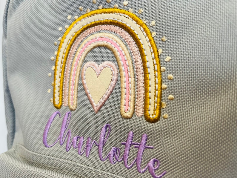 May include: A gray canvas backpack with a rainbow and heart embroidered design. The rainbow is in shades of yellow, pink, and cream. The heart is cream with pink stitching. The name "Charlotte" is embroidered in purple below the rainbow.