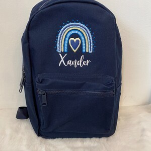 May include: A navy blue canvas backpack with a rainbow and heart design embroidered on the front. The name "Xander" is embroidered below the design.