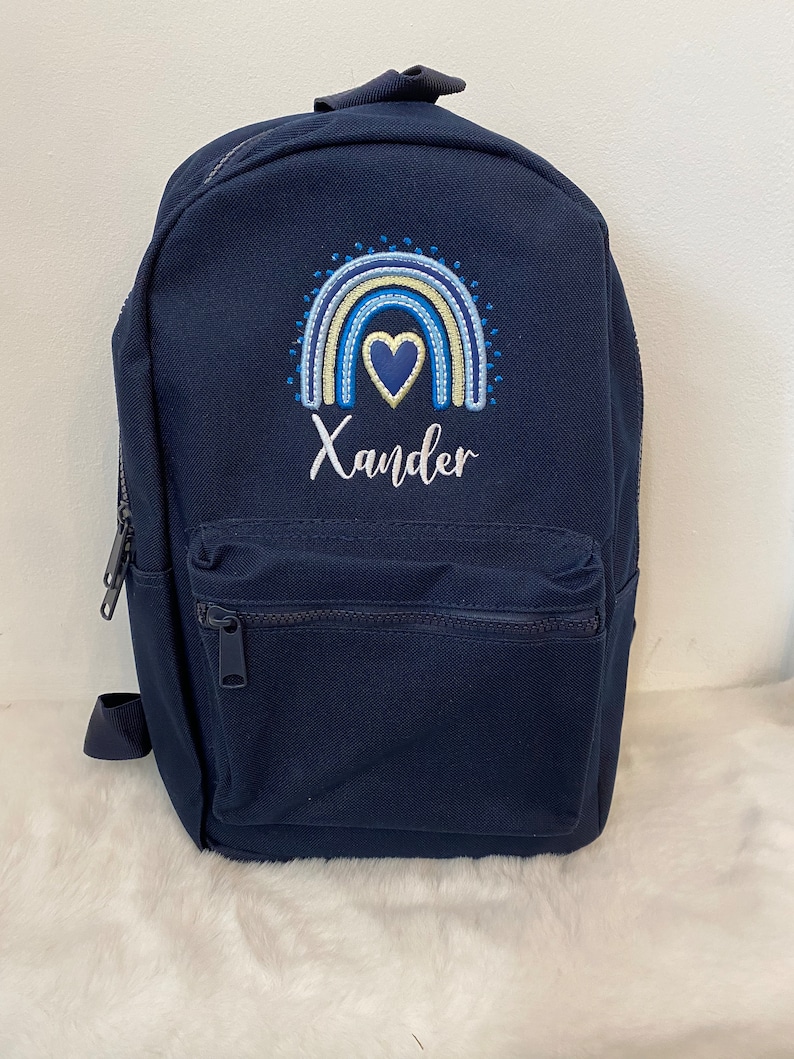 May include: A navy blue canvas backpack with a rainbow and heart design embroidered on the front. The name "Xander" is embroidered below the design.