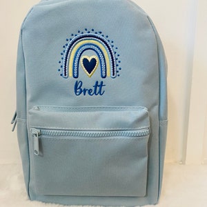May include: Light blue backpack with a rainbow and heart embroidery. The name "Brett" is embroidered below the rainbow.