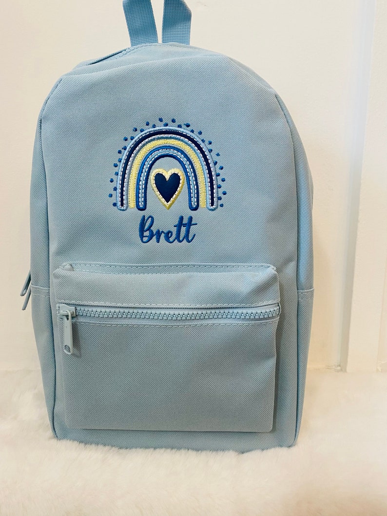 May include: Light blue backpack with a rainbow and heart embroidery. The name "Brett" is embroidered below the rainbow.