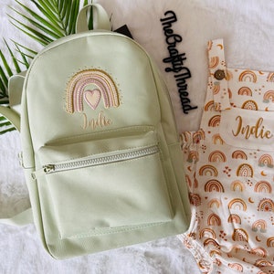 May include: A light green canvas backpack with a rainbow and heart embroidery and the name "Indie" embroidered below. A white onesie with a rainbow and heart pattern and the name "Indie" embroidered on the front pocket.