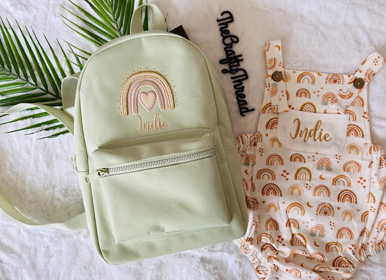 May include: A light green canvas backpack with a rainbow and heart embroidery and the name "Indie" embroidered below. A white onesie with a rainbow and heart pattern and the name "Indie" embroidered on the front pocket.