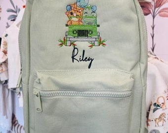 May include: A light green canvas backpack with a zippered front pocket. The backpack has an embroidered design of three dinosaurs riding in a jeep. The name "Riley" is embroidered below the design.
