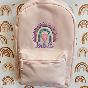 May include: A pink backpack with a rainbow and heart embroidered design. The backpack has a zippered front pocket and the name "Isabelle" is embroidered below the rainbow.