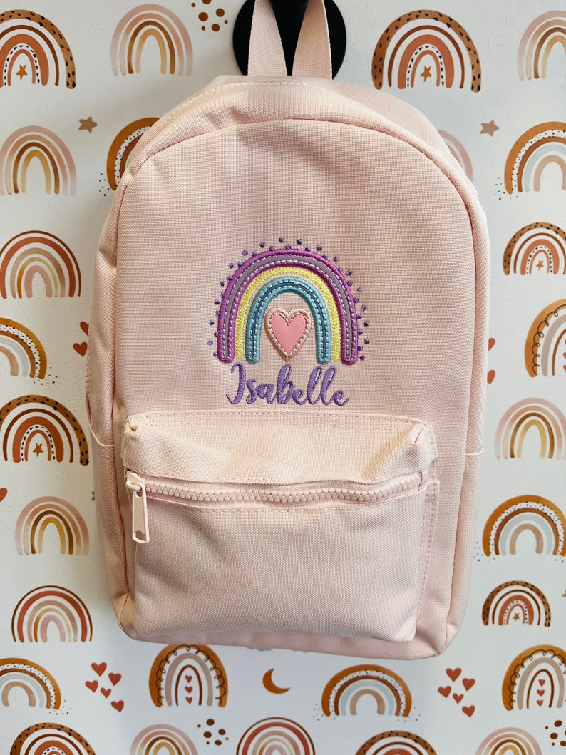 May include: A pink backpack with a rainbow and heart embroidered design. The backpack has a zippered front pocket and the name "Isabelle" is embroidered below the rainbow.