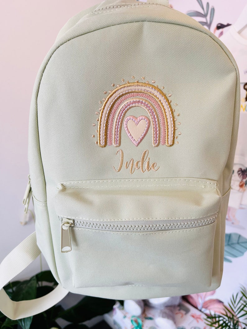May include: A light green canvas backpack with a rainbow and heart embroidery. The name "Indie" is embroidered below the rainbow.