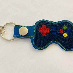 May include: A blue leather keychain shaped like a video game controller with green stitching. The controller has red, yellow, and blue buttons.