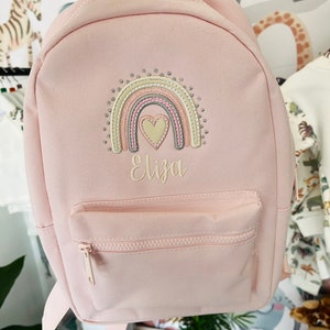 May include: A pink canvas backpack with a rainbow and heart embroidery. The backpack has a front pocket with a zipper and the name "Eliza" embroidered below the rainbow.