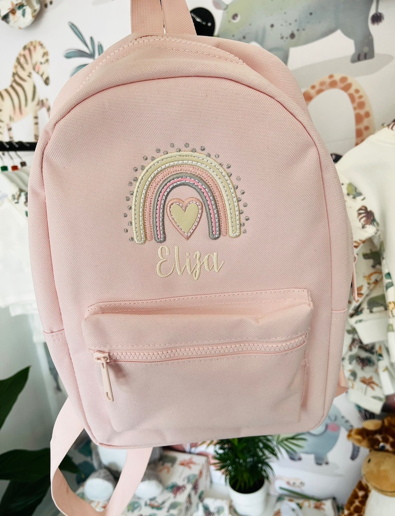 May include: A pink canvas backpack with a rainbow and heart embroidery. The backpack has a front pocket with a zipper and the name "Eliza" embroidered below the rainbow.