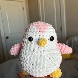 May include: A crocheted penguin plush toy with a pink and white body, yellow beak, and black eyes.
