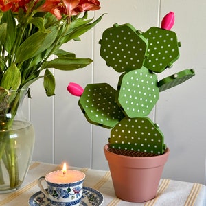 Puede incluir: Un cactus verde y blanco con lunares y flores rosas en una maceta rosa. El cactus está hecho de madera y está sentado sobre un mantel a rayas blancas y amarillas. Hay una vela encendida en una taza de té en la mesa.