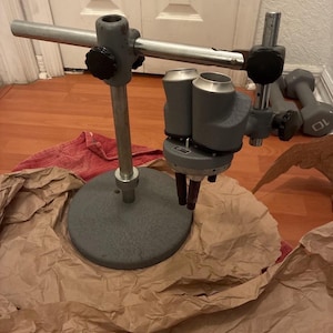 May include: A grey industrial microscope stand with a circular base and adjustable arm. The stand has a vertical pole and a horizontal arm with a black knob. The microscope head has two cylindrical components. The stand is on brown paper.