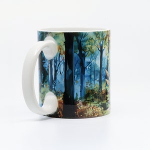 May include: White ceramic mug with a blue and green watercolor design of a forest scene. The mug has a large handle.