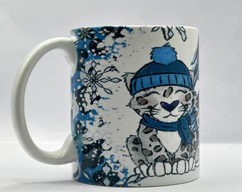 May include: White ceramic mug with a blue and white winter scene featuring a snow leopard wearing a blue hat and scarf. The mug has a white handle.