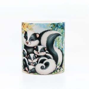 May include: A white ceramic mug with a colorful illustration of a mother skunk and two baby skunks. The skunks are surrounded by green leaves and flowers.