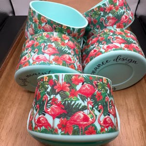 May include: Set of six turquoise blue ceramic bowls with a tropical flamingo and floral design. The bowls are stacked on top of each other. The bowls have a white interior and a black handwritten inscription that reads "High Maintenance Design".