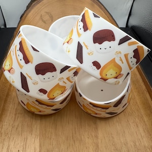 May include: A stack of four white ceramic bowls with a repeating pattern of cartoon marshmallows, chocolate bars, and campfires. The bowls are arranged on a wooden tray, showcasing the design.
