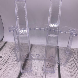 May include: Clear plastic test tube rack with two tall, rectangular test tube holders. The rack has a curved base with multiple small holes for additional test tubes. The background is a light gray.
