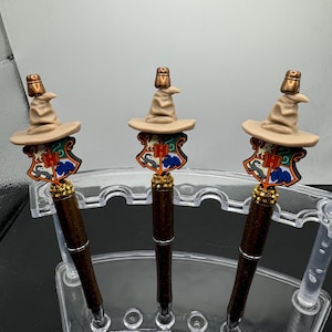 May include: Three brown pens with a Sorting Hat and Hogwarts house crest on top. The pens are displayed on a clear plastic stand.