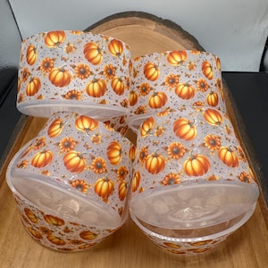 May include: Four clear, cylindrical containers with a fall-themed design. The containers are decorated with orange pumpkins and yellow sunflowers on a light gray background. The containers are arranged on a wooden tray.