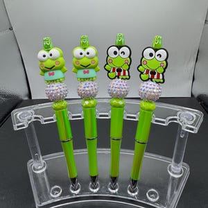 May include: Four green pens with a green frog character on top. The frog characters are wearing different outfits. The pens are displayed in a clear plastic stand.