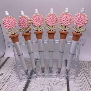 May include: Set of six white pens with pink and beige flower toppers. The pens are displayed in a clear plastic holder.