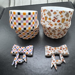 May include: Two cylindrical silicone bands and two bow-shaped accessories. One band and bow feature a multi-colored checkerboard pattern in orange, purple, black, and white. The other band and bow display a pattern of iced coffee cups and pumpkins.