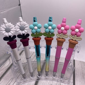 May include: Six pens with flower-shaped tops in a clear plastic holder. The flowers are white, blue, and pink. The pens are silver, gold, and pink.