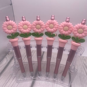 May include: Set of six pink flower pens with a rose gold finish. The pens are arranged in a clear plastic holder.