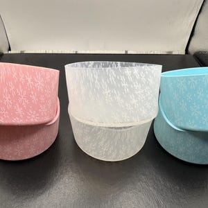 May include: Three decorative hat boxes in pastel colors: pink, white, and blue. Each box features a snowflake pattern. The boxes are stacked and nested, showcasing their varying sizes and designs.