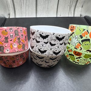 May include: Three round bowls featuring Halloween patterns. The left bowl is pink with skulls, cats, and pumpkins. The center bowl is white with black bats and the word Spooky. The right bowl is light green with ghosts, pumpkins, and Frankenstein faces.