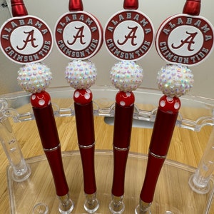 May include: Four red and white pens with a white and iridescent bead and a red and white polka dot bead. Each pen has a round, white and red sticker with the words "Alabama Crimson Tide" and a white "A" in the center.