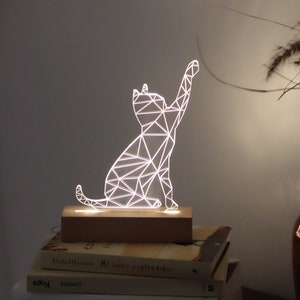 May include: A white geometric cat-shaped night light on a wooden base. The light is on and illuminating the cat shape.