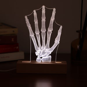 Op de afbeelding: Een wit skeletvormig handvormig nachtlampje met een houten voet. Het licht is aan en verlicht de hand.