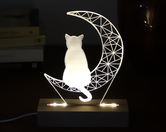 May include: A white cat silhouette sitting on a geometric crescent moon nightlight. The nightlight is made of clear acrylic and has a wooden base.