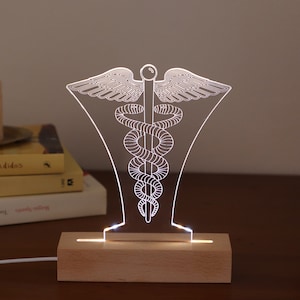 Può includere: Una luce notturna in acrilico bianco illuminata a forma di simbolo medico, il bastone di Asclepio, con una base in legno.